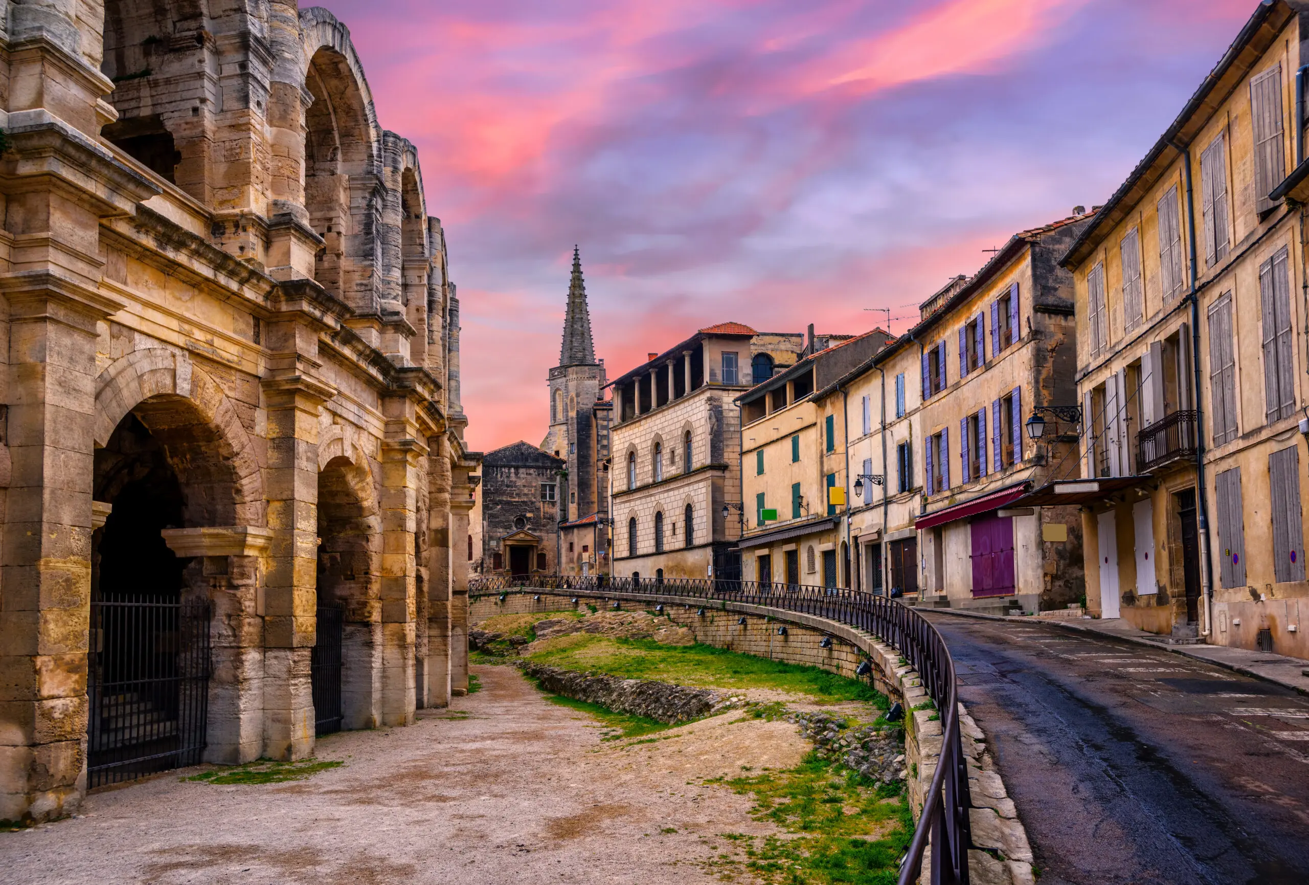 Arles Amphitheatre and Old Town at Dusk, France Stone arches of the Arles Amphitheatre beside a curved old-town street and shuttered buildings beneath a pink evening sky.