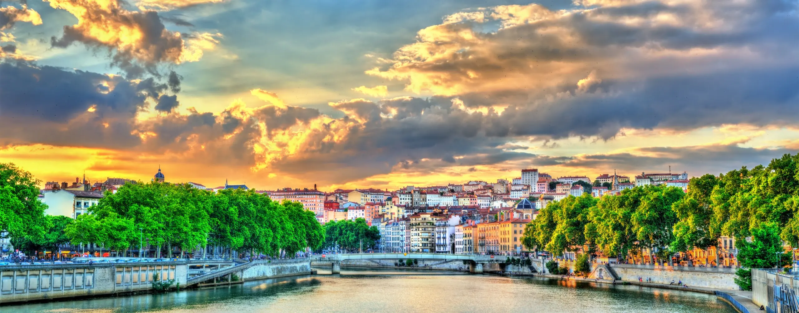 Sunset above the Saone river in Lyon, France Stone arches of the Arles Amphitheatre beside a curved old-town street and shuttered buildings beneath a pink evening sky.