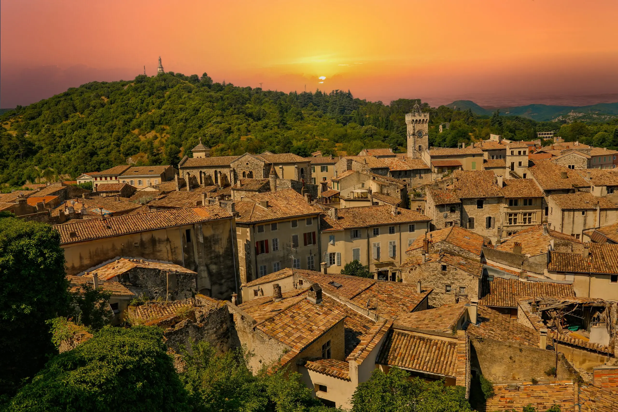 Viviers Old Town at Sunset, France Terracotta rooftops and a stone clock tower in Viviers, France, beneath an orange sunset sky with wooded hills beyond.